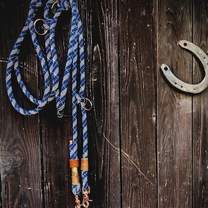 May include: A blue and white braided rope leash featuring metallic hardware, including circular rings and two swivel snap hooks at the ends, accented with tan leather wraps and gold-toned wire detailing near the bottom attachment points.