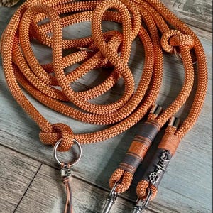 Pode incluir: Trela para cães de corda laranja com fechos e detalhes em metal prateado. A trela é enrolada e apresenta um nó e uma borla decorativa. As pegas são envolvidas em tecido cinzento e laranja.