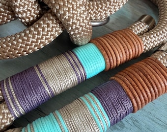 May include: A braided rope leash featuring a metallic champagne-colored cord with decorative wraps in brown, purple, and turquoise thread. The leash includes a metal ring and a cylindrical metal end cap, showcasing a multi-colored woven design.