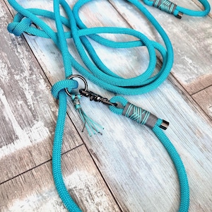 May include: A turquoise dog leash and matching collar set. The leash is made of thick, braided rope with a metal clasp and decorative gray and turquoise accents. The collar has a similar design. The set is displayed on a wooden surface.