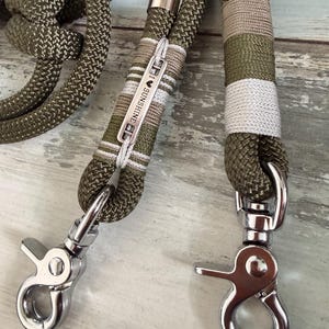 May include: An olive green braided rope leash featuring two silver-toned metal swivel snap hooks. The leash includes decorative wrapping in olive, beige, and white thread, with a small metal tag engraved with the text I HEART SUNSHINE.
