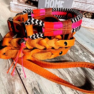 May include: A stack of dog collars and a matching lead. The collars are made of brown leather and feature decorative woven and braided accents in black, white, pink, and orange. The lead is orange.