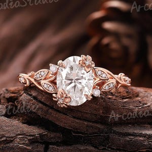 May include: Rose gold engagement ring with an oval-cut diamond center stone. The ring features floral and leaf accents with smaller diamonds. The ring is set on a dark brown wooden surface.