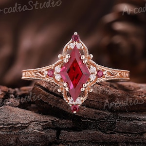 May include: A rose gold ring with a large, red, diamond-shaped center stone. The ring features smaller red and white stones, and intricate filigree detailing. The ring is set against a dark, textured background.