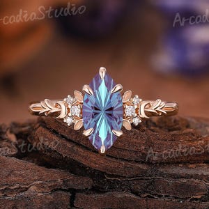 May include: A gold-toned ring with a marquise-cut, blue-green gemstone at the center. The ring features floral and leaf accents with small, clear gemstones. The ring is set against a textured, brown background.