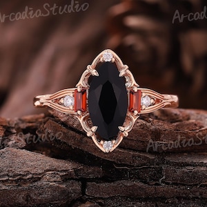 May include: A rose gold ring featuring a large, oval-cut black gemstone at the center, flanked by two rectangular red gemstones. The ring has a decorative filigree design and small white accent stones.