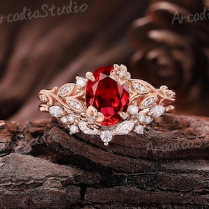 May include: A rose gold ring featuring an oval, deep red gemstone as the centerpiece. The ring is adorned with floral and leaf designs, and small white gemstones. The ring is set against a dark, textured background.