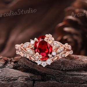 May include: Rose gold ring featuring a large oval-cut red gemstone, accented with small white gemstones and floral and leaf detailing. The ring is set against a natural wood background, showcasing its intricate design.