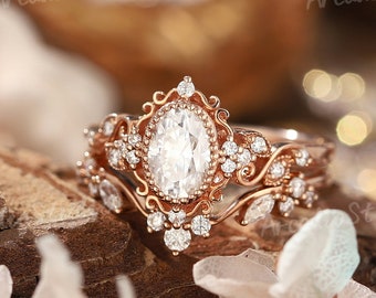 Könnte beinhalten: Ein Ring aus Roségold mit einem zentralen, oval geschliffenen weißen Stein, umgeben von einer dekorativen Filigranfassung mit kleinen runden Akzentsteinen. Die Ringschiene zeigt filigrane Schnörkel und weitere kleine Steine.