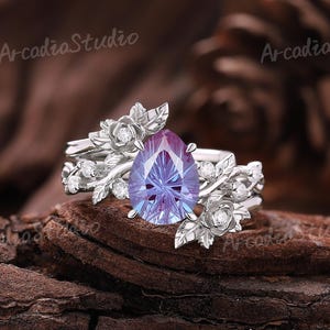May include: A silver ring with a pear-shaped, lavender-colored gemstone. The ring features floral and leaf accents with small, clear stones. The words "Arcadia Studio" are printed on the top left and right of the image.