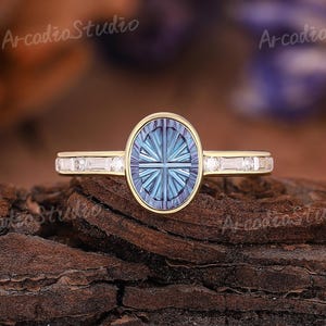 May include: A gold ring featuring an oval-shaped blue gemstone with a faceted sunburst pattern in a bezel setting. The band is accented with small, clear baguette-cut stones on each side. The text ArcadiaStudio is visible in the background.
