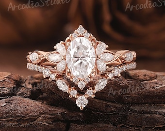 Puede incluir: Un anillo de compromiso de oro rosa con una piedra central ovalada y una banda delicada con piedras más pequeñas. El anillo está colocado sobre una superficie de madera.
