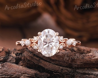 May include: A rose gold engagement ring with an oval-shaped center stone and small round stones on the band. The ring also features two pearl accents on each side of the center stone.