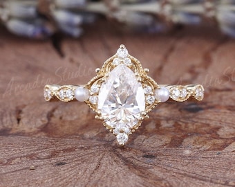 May include: A gold engagement ring with a pear-shaped diamond center stone. The ring features a halo of smaller diamonds and pearls, with intricate detailing on the band. The ring is set against a wooden surface with a blurred background.