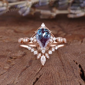 Könnte beinhalten: Ein birnenförmiger Alexandrit-Verlobungsring in Roségold mit einem Halo aus kleineren Diamanten. Der Ring ist mit einem zarten Band aus Diamanten verziert.