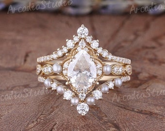 May include: A pear-shaped diamond engagement ring set with a delicate band and a halo of smaller diamonds. The ring is accented with pearls and has a vintage-inspired design.