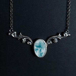 May include: A silver-toned necklace with an oval pendant featuring a blue and white floral design. The pendant is framed by a silver setting and connected to a delicate chain. The necklace has ornate, curved details on either side of the pendant.
