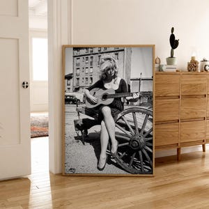 Pu&ograve; includere: Una stampa artistica da parete incorniciata in bianco e nero che ritrae una persona con capelli alle spalle seduta su una ruota di carro in legno mentre suona una chitarra acustica. Lo sfondo mostra un edificio e un'auto parcheggiata.