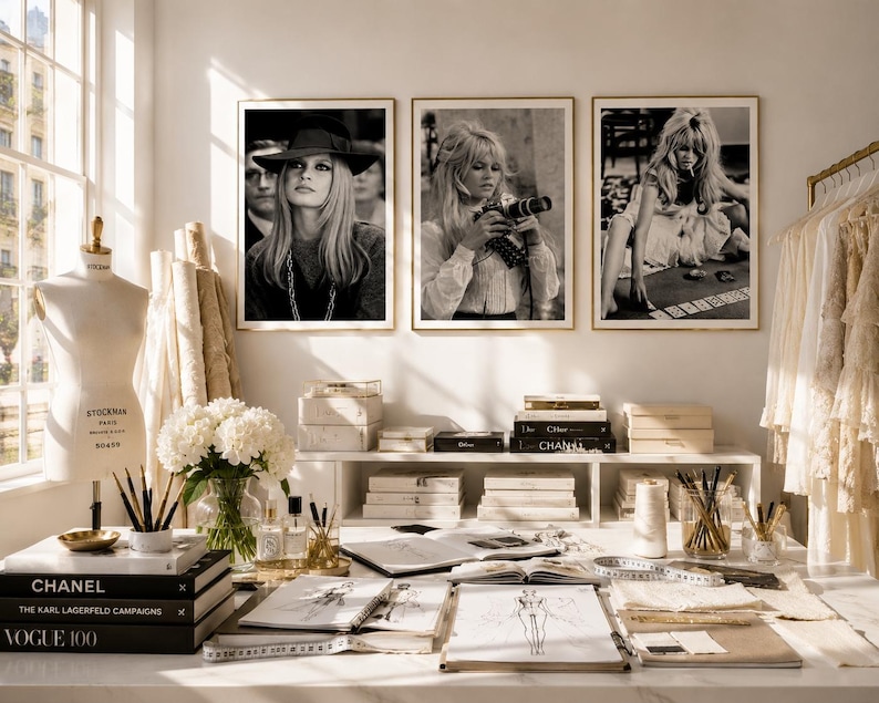 Op de afbeelding: Een mode-ontwerpstudio met drie ingelijste zwart-witfoto's van een vrouw. De werkruimte omvat een paspop, ontwerpschetsen, boeken en een kledingrek. De boeken bevatten "CHANEL" en "VOGUE 100".