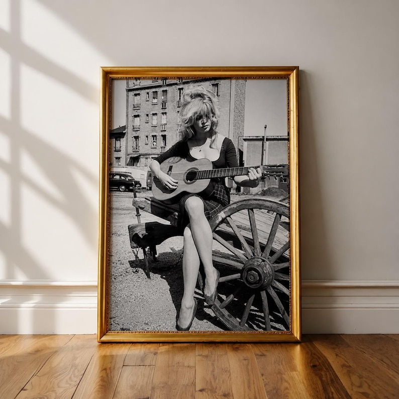 Pu&ograve; includere: Una fotografia incorniciata in bianco e nero che ritrae una persona con capelli voluminosi seduta su una ruota di carro in legno mentre suona una chitarra acustica. La persona indossa un top scuro a maniche lunghe e una gonna all'aperto.