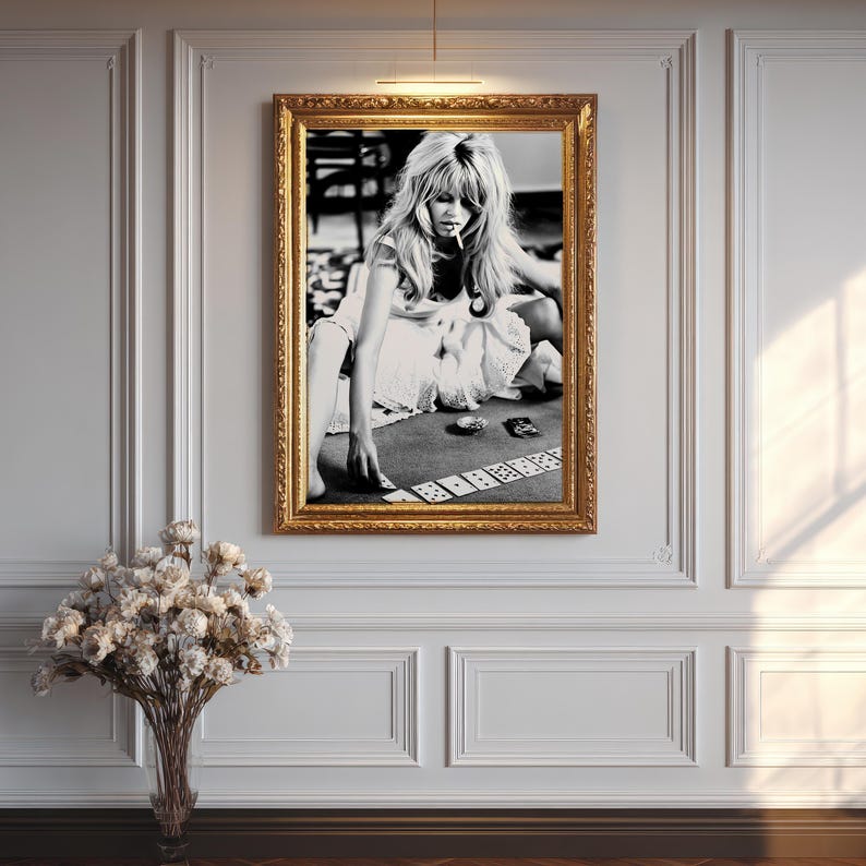 Op de afbeelding: Zwart-wit ingelijste foto van een vrouw die kaarten speelt. De foto staat in een gouden, sierlijke lijst en hangt aan een witte muur. Een boeket gedroogde bloemen staat in een glazen vaas onder de foto.