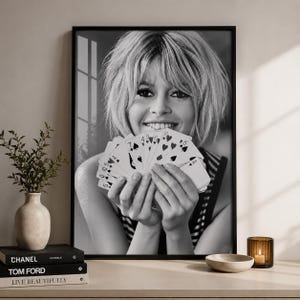 K&ouml;nnte beinhalten: Ein schwarz-wei&szlig; gerahmter Wandkunstdruck, der das Portr&auml;t einer Person mit kurzem Haar zeigt, die einen F&auml;cher aus Spielkarten vor das Gesicht h&auml;lt. Die Person l&auml;chelt und die Karten zeigen deutlich verschiedene Symbole und Zahlenwerte.