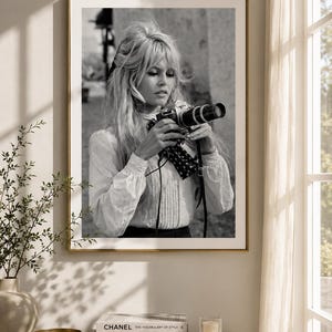 Op de afbeelding: Zwart-wit ingelijste foto van een vrouw met een camera. De vrouw draagt een witte blouse en heeft blond haar. De foto wordt aan een muur getoond boven een marmeren kast met boeken en een kaars.