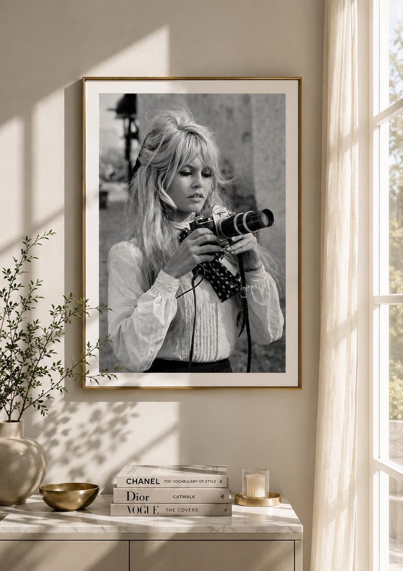 Op de afbeelding: Zwart-wit ingelijste foto van een vrouw met een camera. De vrouw draagt een witte blouse en heeft blond haar. De foto wordt aan een muur getoond boven een marmeren kast met boeken en een kaars.