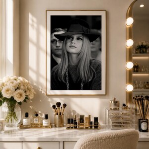 Op de afbeelding: Zwart-witfoto in een lijst van een vrouw met een hoed, met een man op de achtergrond. De foto wordt tentoongesteld boven een kaptafel met make-up en accessoires. Een boeket witte rozen staat in een glazen vaas.