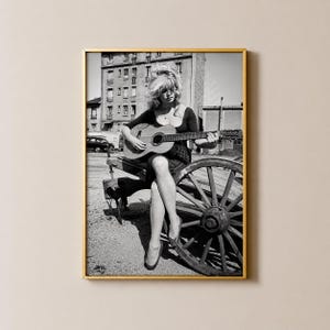 Pu&ograve; includere: Una stampa artistica incorniciata in bianco e nero che ritrae una persona con capelli voluminosi seduta su una ruota di carro in legno, mentre suona una chitarra acustica. Lo sfondo mostra un edificio a pi&ugrave; piani e un'auto d'epoca.