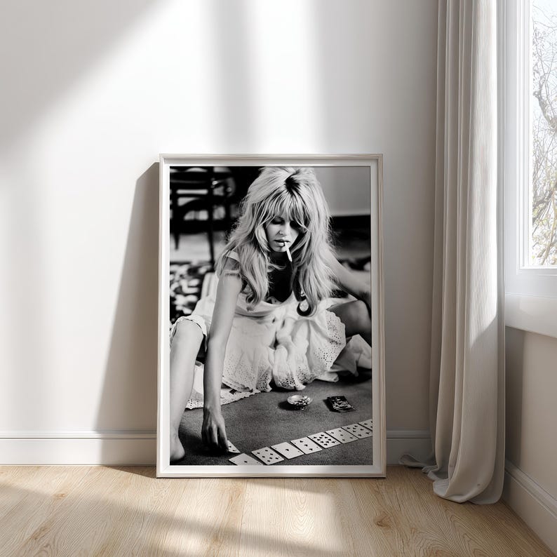 Op de afbeelding: Zwart-wit ingelijste print met een vrouw die kaarten speelt. De vrouw draagt een witte jurk en heeft lang, golvend haar. Ze heeft een sigaret in haar mond. De print wordt weergegeven op een muur naast een raam met witte gordijnen.