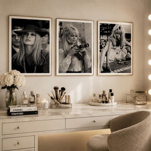 Op de afbeelding: Drie ingelijste zwart-witfoto's van een vrouw hangen boven een kaptafel. De witte kaptafel met gouden hardware bevat parfumflesjes, make-upborstels en boeken. Een verlichte spiegel en een beige stoel maken de sc&egrave;ne compleet.