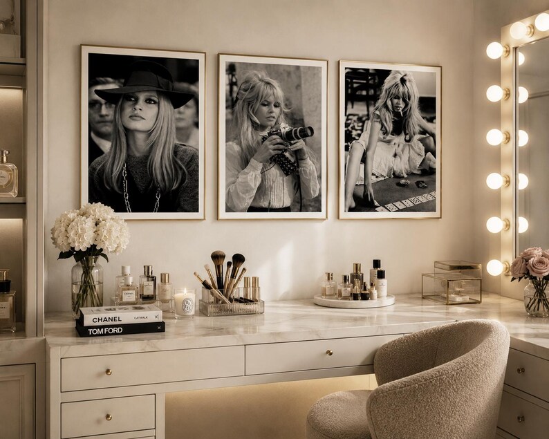 Op de afbeelding: Drie ingelijste zwart-witfoto's van een vrouw hangen boven een kaptafel. De witte kaptafel met gouden hardware bevat parfumflesjes, make-upborstels en boeken. Een verlichte spiegel en een beige stoel maken de sc&egrave;ne compleet.