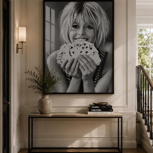 K&ouml;nnte beinhalten: Ein gro&szlig;er, schwarz-wei&szlig; gerahmter Wandkunstdruck, der das Portr&auml;t einer Person mit kurzem Haar zeigt, die einen F&auml;cher aus Spielkarten vor das Gesicht h&auml;lt. Das Kunstwerk ist &uuml;ber einem rechteckigen Konsolentisch mit einer Vase und B&uuml;chern platziert.