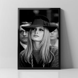 Op de afbeelding: Zwart-wit ingelijste print van een vrouw met een zwarte hoed met brede rand en een strik. Ze heeft lang blond haar en een zilveren ketting. De afbeelding is een portret, met een vage achtergrond.