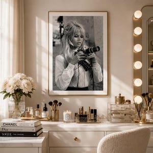Op de afbeelding: Zwart-wit foto in een lijst van een vrouw die een camera vasthoudt. De foto wordt getoond op een witte kaptafel met schoonheidsproducten, boeken en een vaas met bloemen. Rechts een verlichte make-upspiegel.