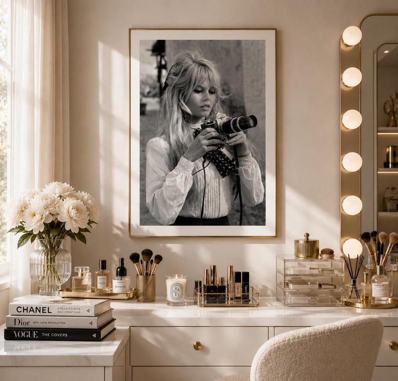 Op de afbeelding: Zwart-wit foto in een lijst van een vrouw die een camera vasthoudt. De foto wordt getoond op een witte kaptafel met schoonheidsproducten, boeken en een vaas met bloemen. Rechts een verlichte make-upspiegel.