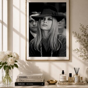 Op de afbeelding: Zwart-wit ingelijste foto van een vrouw met een zwarte hoed met strik. Ze heeft lang blond haar en een ketting. De foto wordt getoond op een witte kast met boeken, een gouden kom en parfumflesjes.