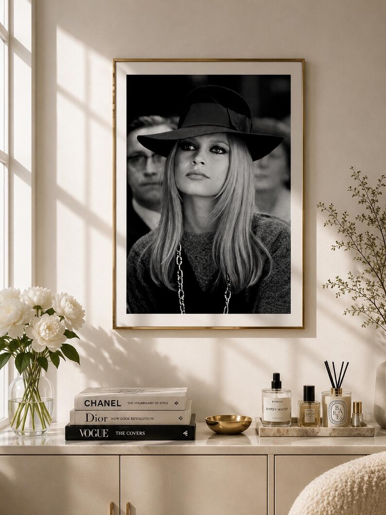 Op de afbeelding: Zwart-wit ingelijste foto van een vrouw met een zwarte hoed met strik. Ze heeft lang blond haar en een ketting. De foto wordt getoond op een witte kast met boeken, een gouden kom en parfumflesjes.
