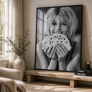 K&ouml;nnte beinhalten: Ein schwarz-wei&szlig; gerahmter Wandkunstdruck, der eine l&auml;chelnde Person mit kurzem Haar zeigt, die einen F&auml;cher aus Spielkarten h&auml;lt. Das Portr&auml;t ist in einem d&uuml;nnen schwarzen Rahmen pr&auml;sentiert und f&auml;ngt einen spontanen Moment ein.
