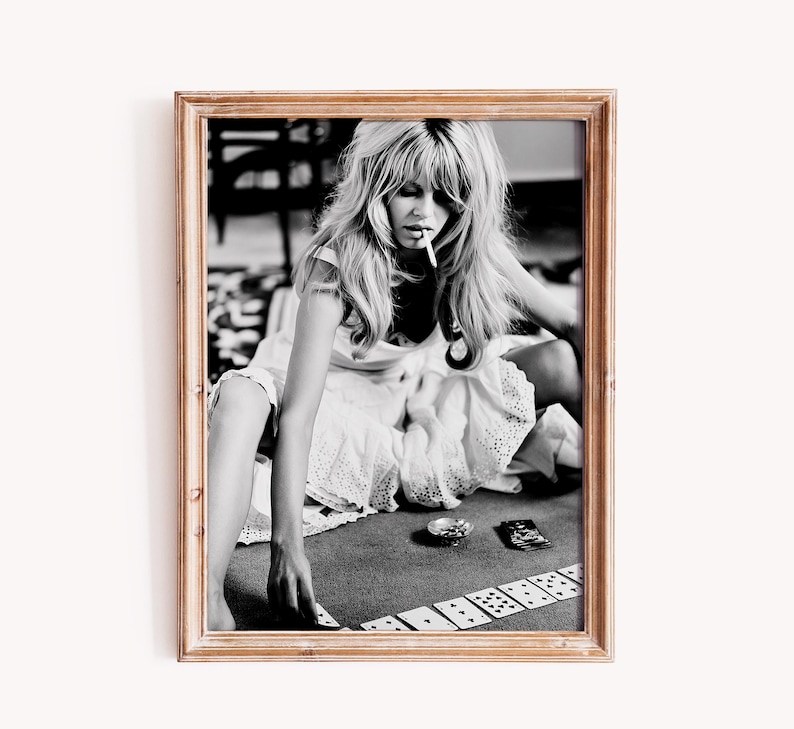 Op de afbeelding: Zwart-wit ingelijste foto van een vrouw met lang blond haar, die een sigaret rookt en kaarten speelt. De vrouw draagt een witte jurk en zit op de grond. De foto zit in een houten lijst.