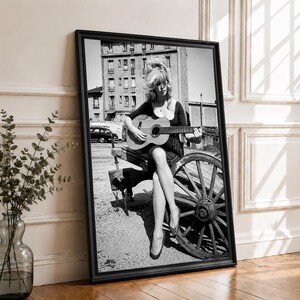 Pu&ograve; includere: Una stampa artistica incorniciata in bianco e nero che ritrae una persona con i capelli lunghi seduta su una ruota di carro in legno mentre suona una chitarra acustica. La scena include un edificio a pi&ugrave; piani sullo sfondo e un'auto parcheggiata.