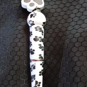 May include: A black and white pen with a paw print design. The pen has a silver metal tip and a black bead with white paw prints at the top.
