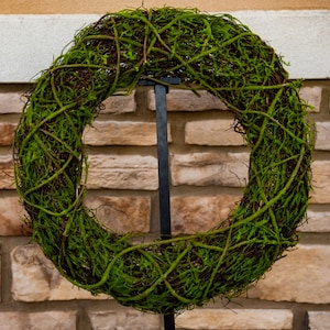 May include: A green moss wreath with brown vines, hanging on a black metal stand against a brick wall.