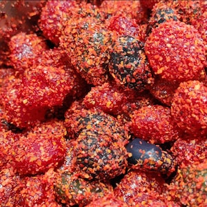 Puede incluir: Primer plano de un montón de caramelos recubiertos de rojo. Los caramelos son una mezcla de colores rojos y oscuros, generosamente cubiertos con copos de chile rojo. La imagen es un primer plano, que muestra la textura y los detalles de los caramelos.