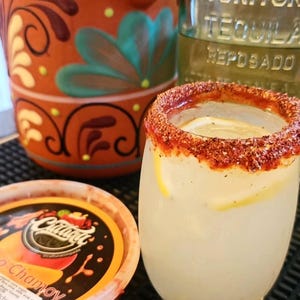 Puede incluir: Un cóctel refrescante en un vaso con borde de chile en polvo, adornado con rodajas de limón. Una jarra de terracota con diseños florales y una botella de tequila están en el fondo. Un recipiente de chamoy está en primer plano.