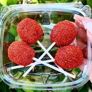 May include: Four red, round lollipops covered in red chili powder are arranged in a clear plastic container. The lollipops are on white sticks.