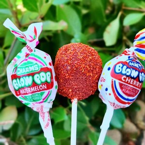 May include: Three lollipops on sticks. The lollipop on the left is a watermelon flavored Blow Pop. The lollipop on the right is a cherry flavored Blow Pop. The lollipop in the middle is a red, round lollipop covered in red sprinkles.