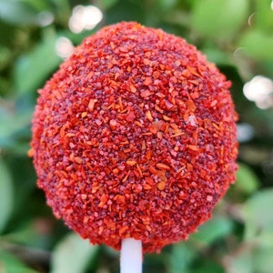 May include: A red cake pop covered in red sprinkles on a white stick. The cake pop is round and has a smooth surface. The sprinkles are small and evenly distributed.