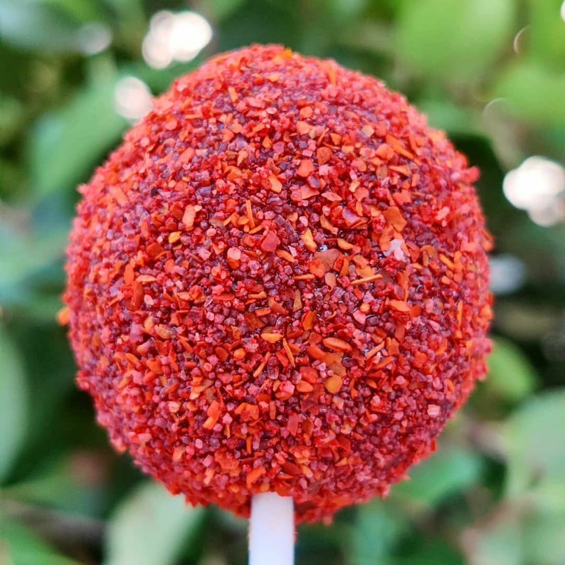 May include: A red cake pop covered in red sprinkles on a white stick. The cake pop is round and has a smooth surface. The sprinkles are small and evenly distributed.